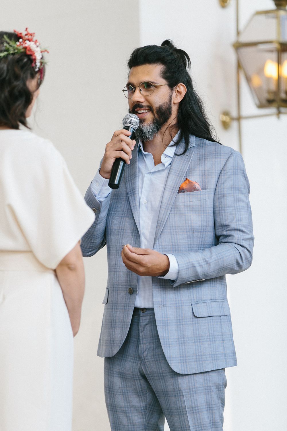 A groom in a light blue checkered suit speaks into a microphone during a wedding ceremony.