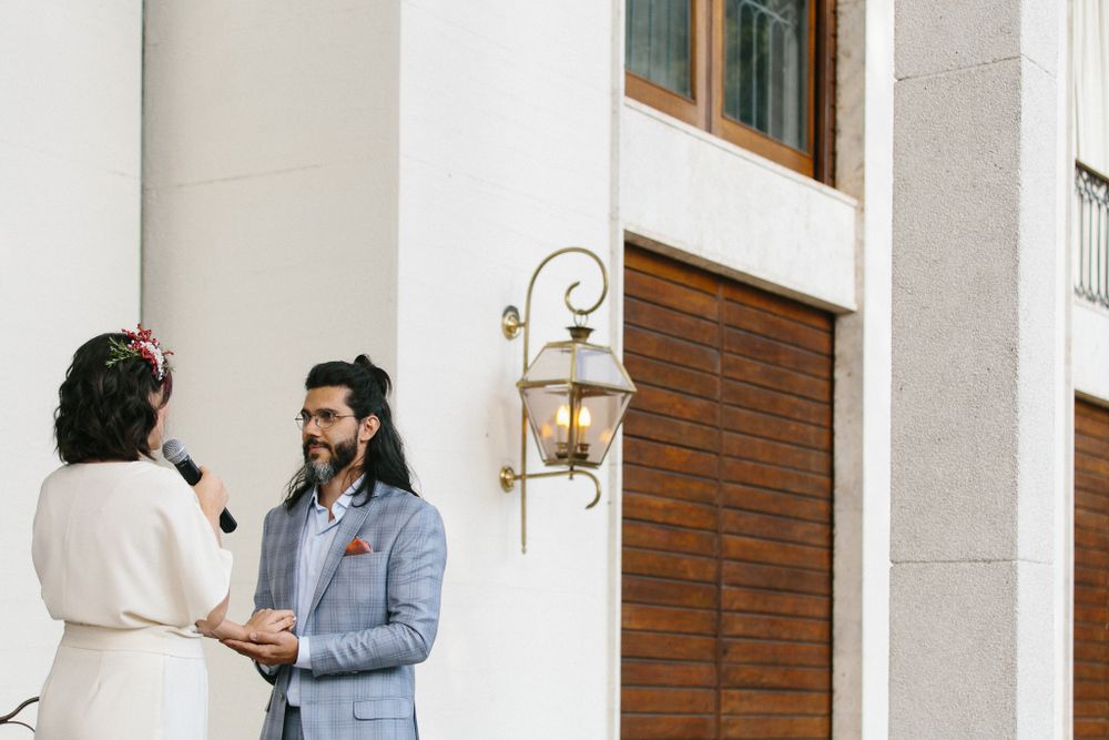 A couple stands intimately close while exchanging vows outside a building with white walls and brick accents.