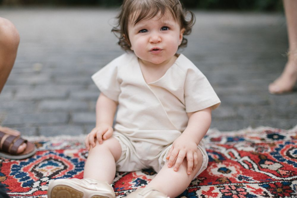 A small baby in cream-colored clothing sits on a colorful patterned rug.