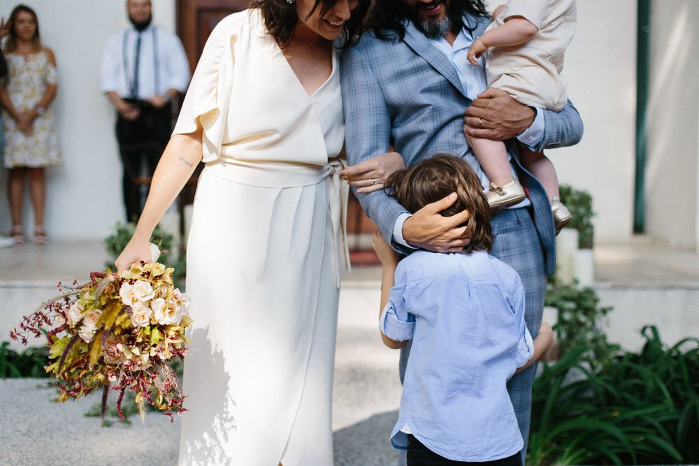 Wedding party gathered outside with children and floral decorations visible in the background.
