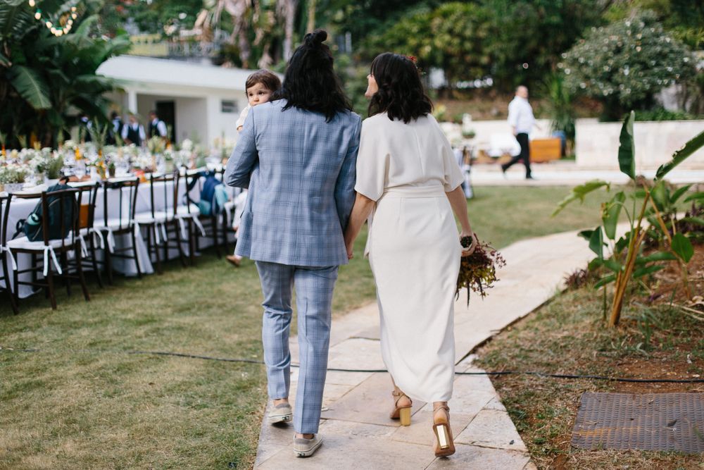 Newlyweds walking along a garden path with outdoor reception tables visible in the background.