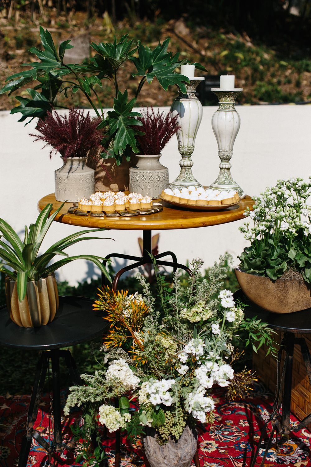 Tiered display of appetizers and decorative glass bottles surrounded by potted plants and flowers.