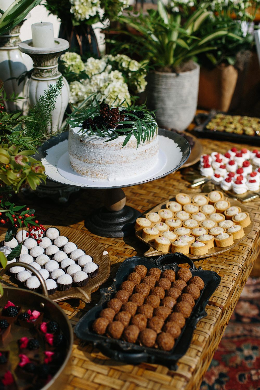 Elegant dessert table with white cake, truffles and various sweet treats surrounded by greenery.