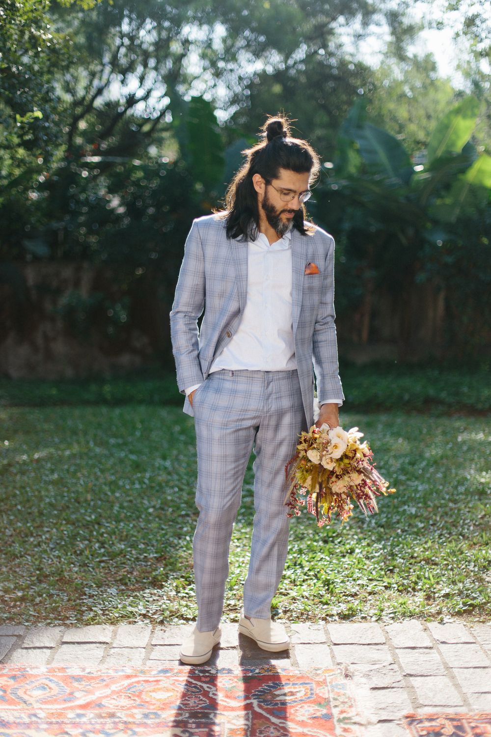 A person in a light gray suit with long hair holding dried flowers stands on a patterned carpet outdoors.