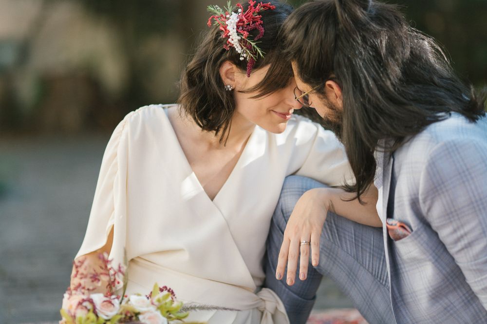 Two people in wedding attire share an intimate moment on a patterned rug in a garden setting.