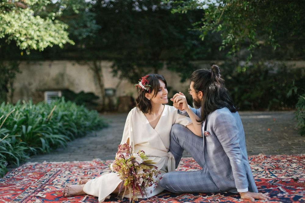 A couple sits together on a decorative rug outdoors with floral arrangements nearby.