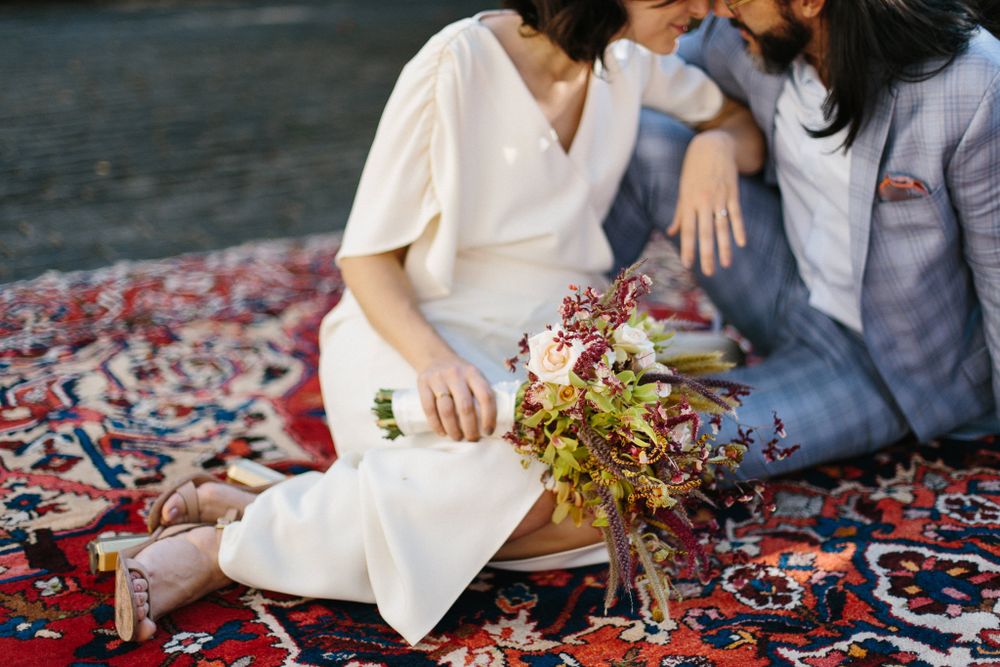 Two people sit on a patterned rug with dried flower bouquets in an outdoor setting.