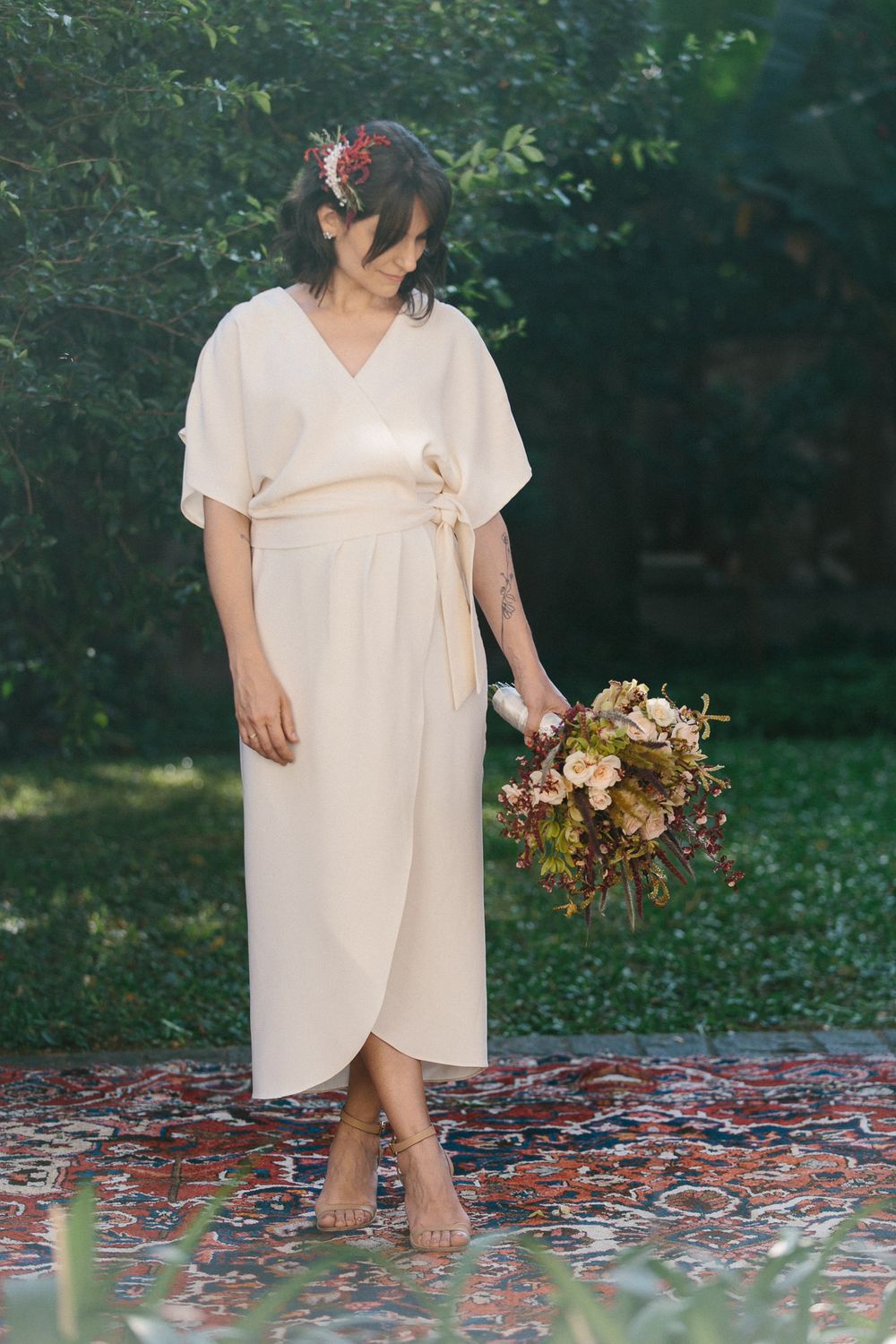 Person in flowing white wrap dress stands barefoot on patterned rug holding dried flowers.