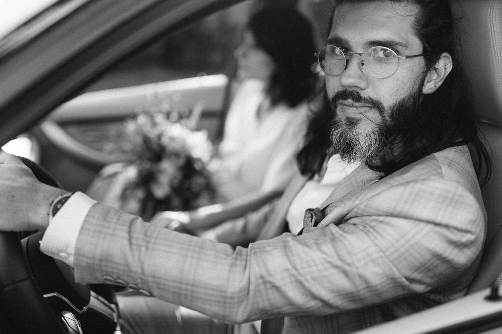 Black and white portrait inside a car showing someone with a beard wearing glasses.