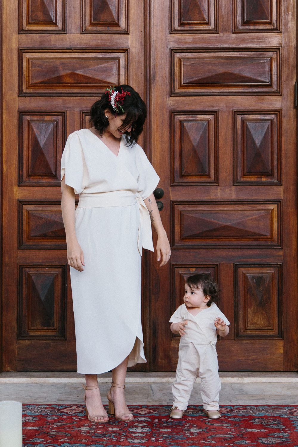 Parent and toddler standing against ornate wooden doors, both wearing light-colored outfits.