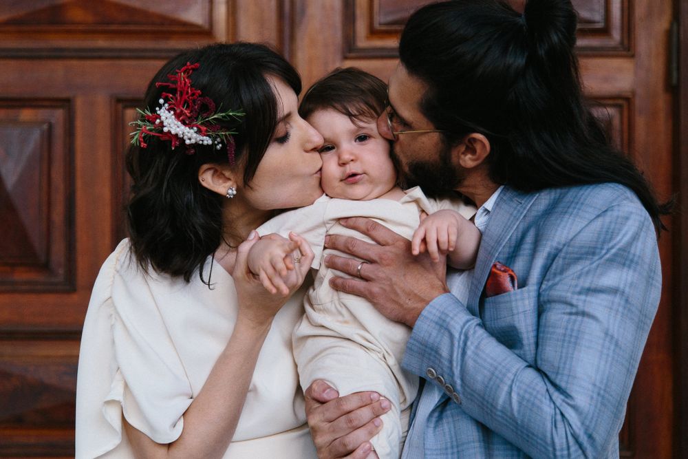 Intimate family moment showing affectionate parents kissing their baby in front of wooden paneled wall.