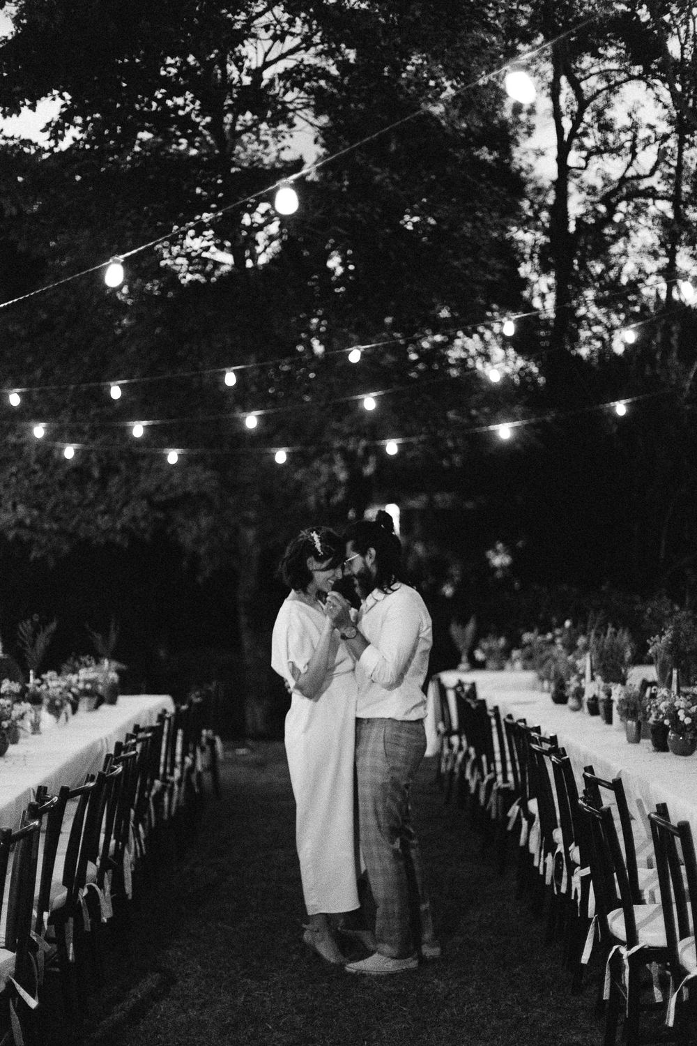 Romantic evening wedding scene with string lights and couple standing between rows of chairs.