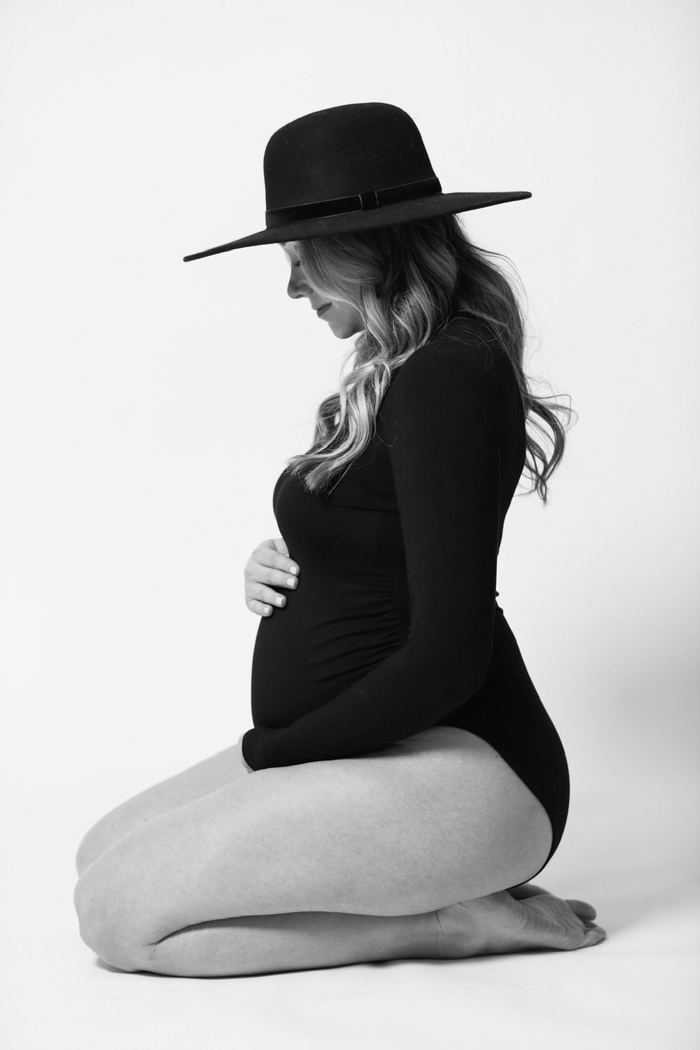 Elegant maternity portrait in black bodysuit and wide-brim hat against white background.