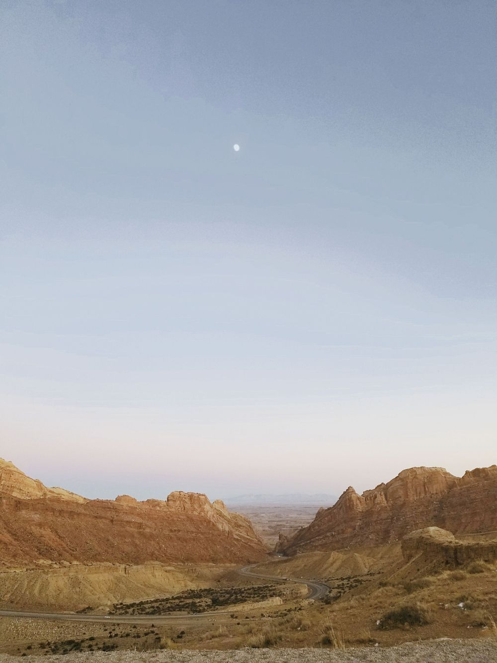 Moon visible over desert canyon landscape with rocky mountains at dusk.