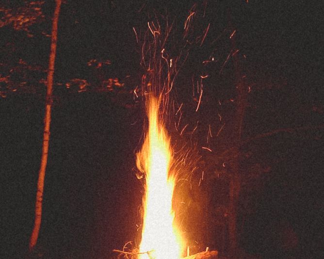 Bright orange campfire flame shooting upward with sparks against dark night sky.