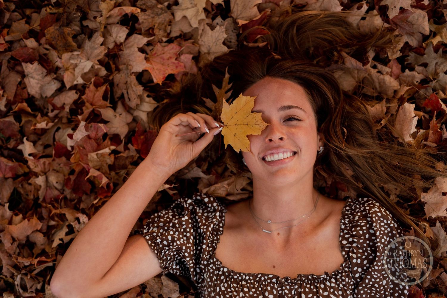 Someone lying in fall leaves holding a yellow maple leaf with a joyful expression.