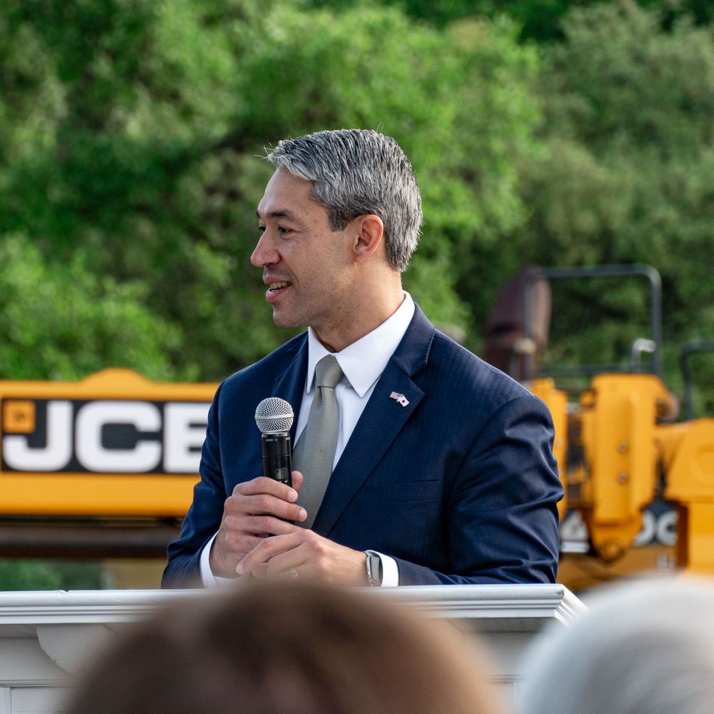Outdoor speaking event with podium and yellow construction equipment visible in background.
