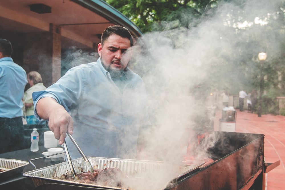 Outdoor barbecue event with smoke rising from grills as people prepare food.
