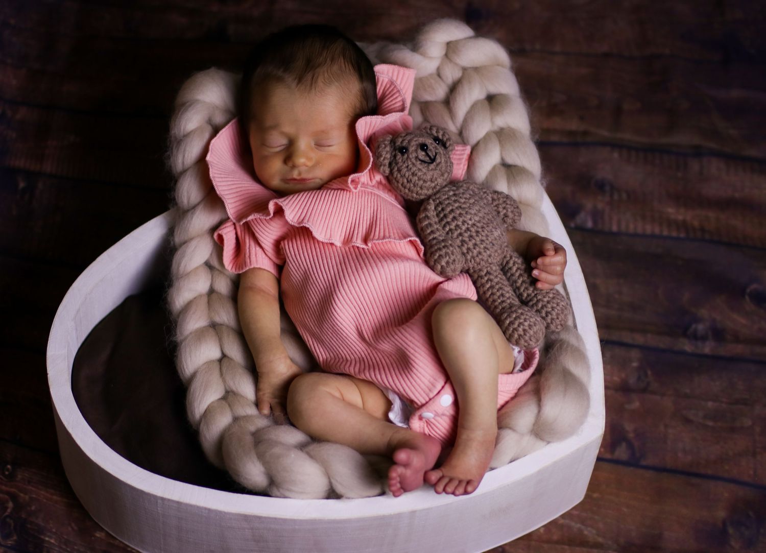 Sleeping newborn in pink onesie with stuffed toy in white heart-shaped basket.