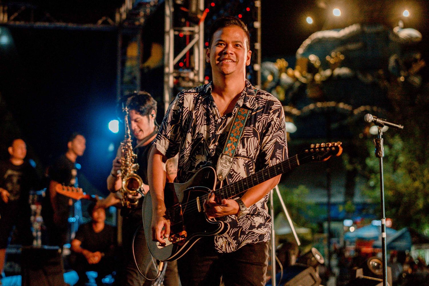 Musician performs on stage wearing a patterned shirt while playing guitar during a nighttime concert.