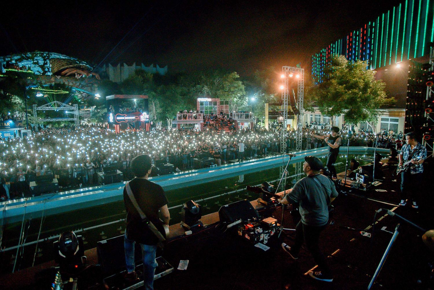 Aerial view of massive concert crowd with stage and city lights illuminating the nighttime scene.