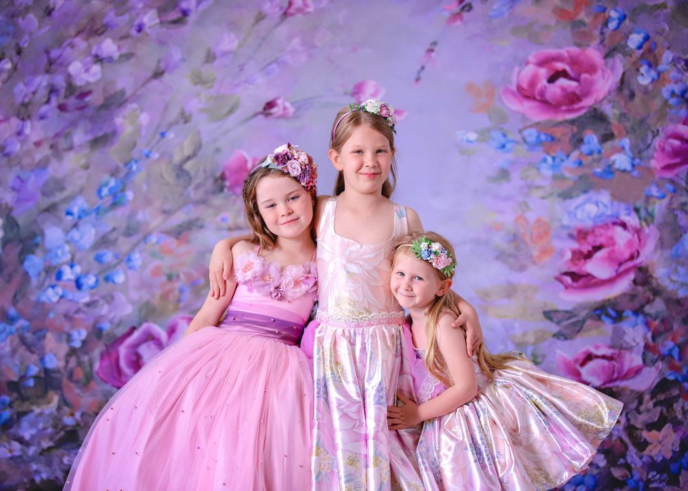 Three girls wearing couture gowns embracing in front of floral studio backdrop for a Dream Dress Session.