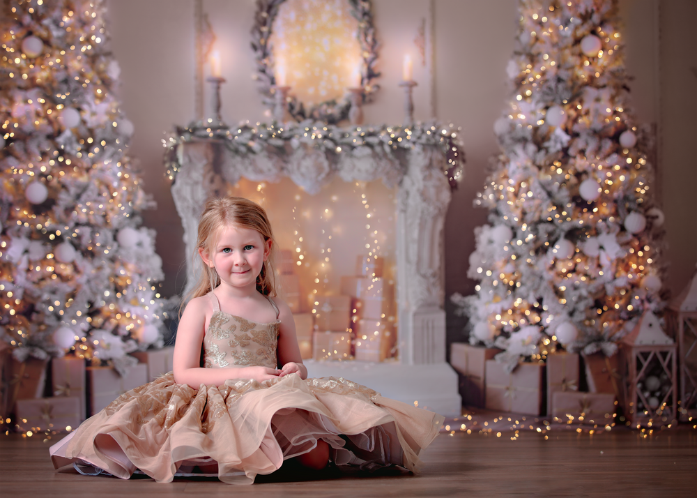 Little girl at Dream Dress Session wearing short gold sequin gown sitting in front of twinkling gold Christmas scene.