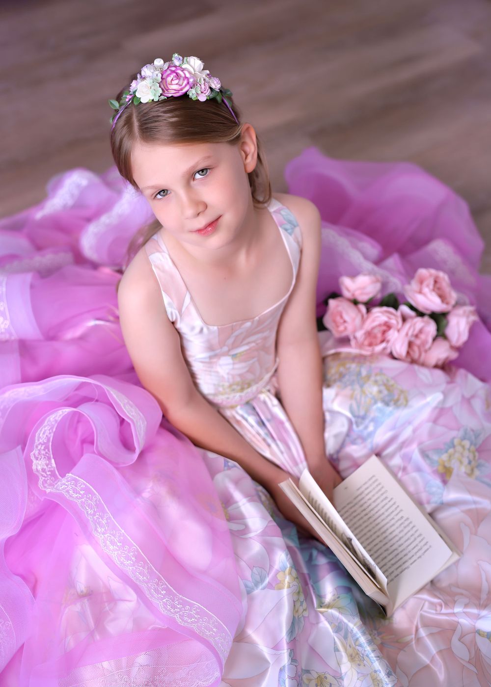 Girl wearing satin couture gown sitting on wooden floor holding a book surrounded by ruffles for a Dream Dress Session.