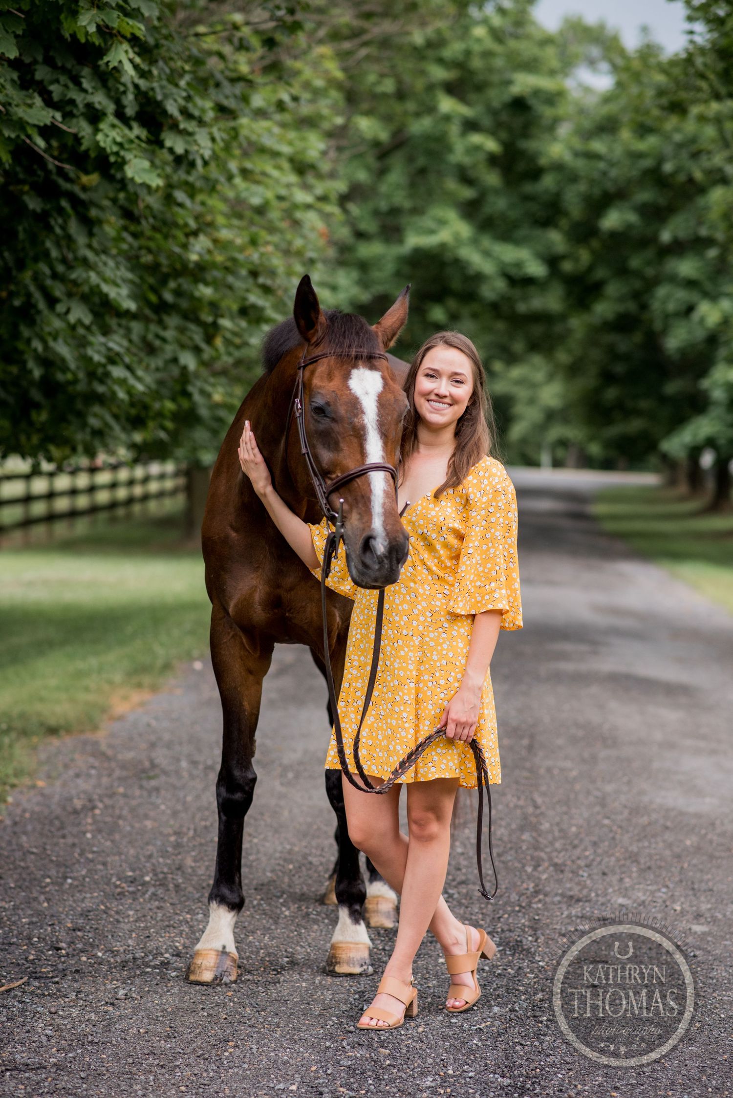 A bay horse and person in yellow dress pose together on a tree-lined path.