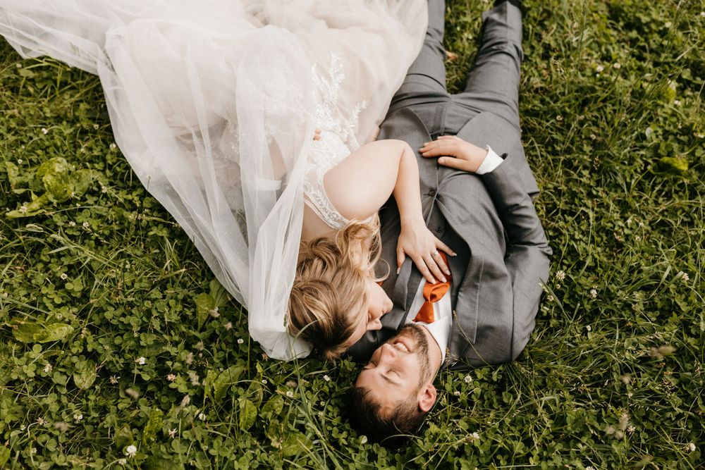 Aerial view of bride in tulle gown and groom in gray suit lying together on green grass.