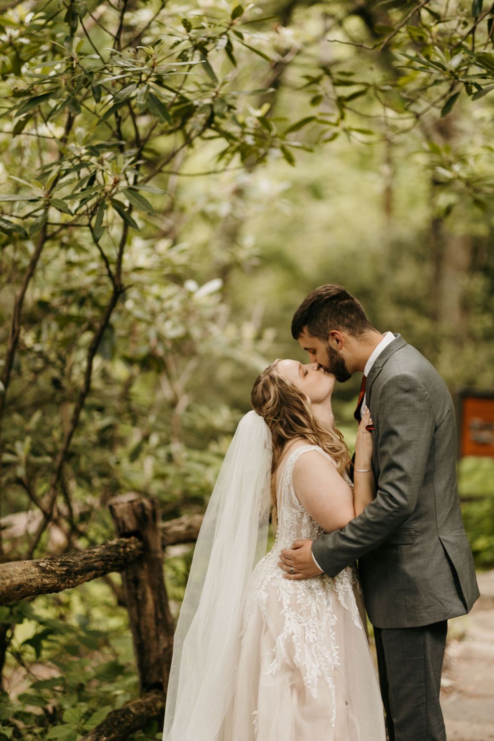 Bride and groom share a romantic kiss surrounded by lush green trees in a forest wedding setting.