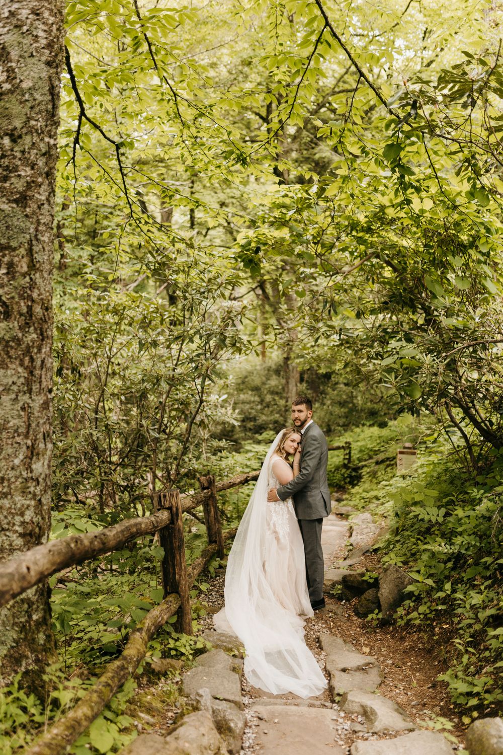 Groom embraces bride in lace gown on a stone forest path surrounded by lush green trees.