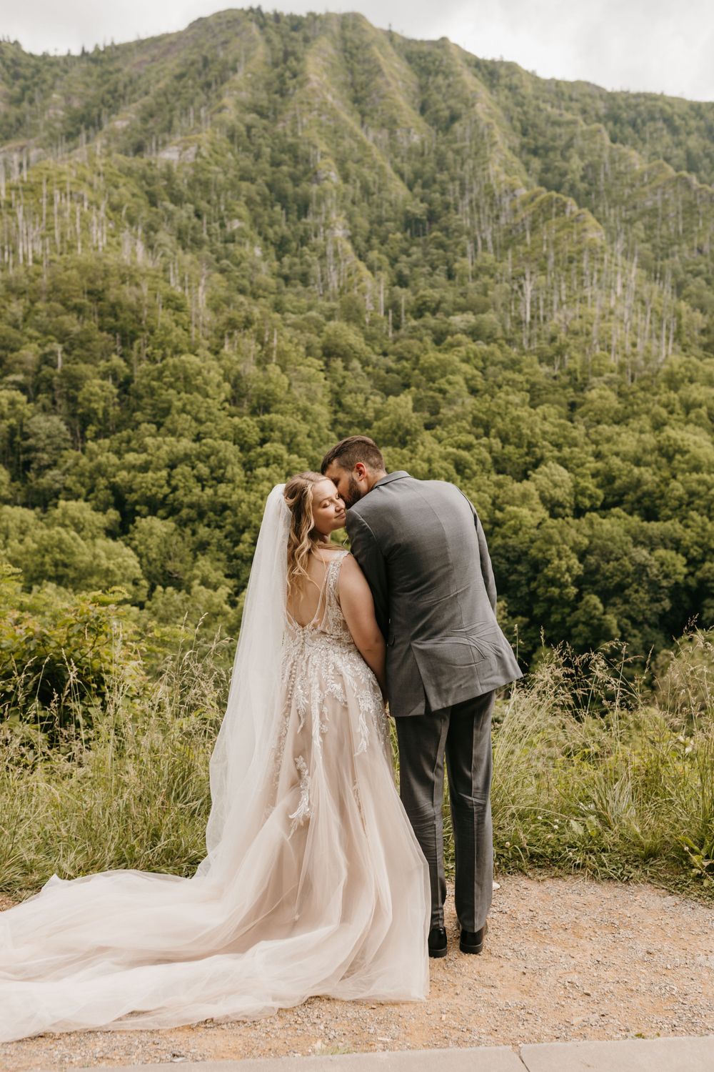 Bride and groom share romantic forehead touch in front of green mountain backdrop on their wedding day.