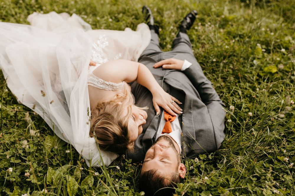 Bride and groom laugh together while lying on green grass in an intimate wedding portrait.