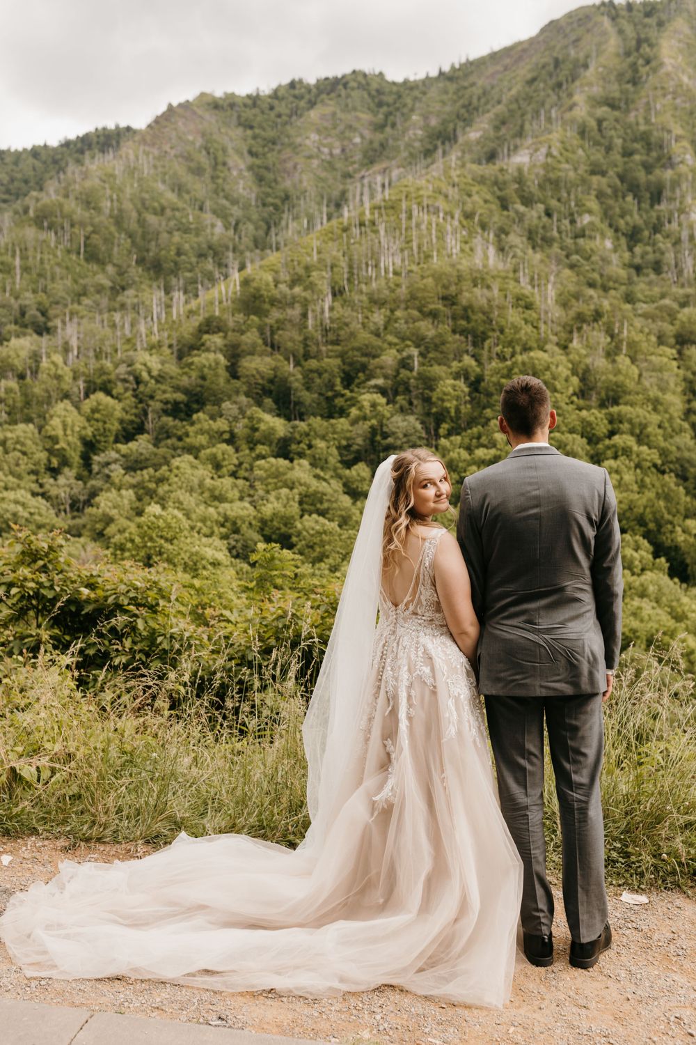 Bride in lace gown with long veil stands with groom in gray suit overlooking lush green mountain landscape.