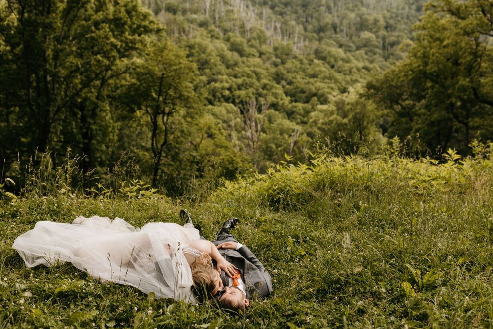 Bride and groom lie together in a sunlit green meadow with forested mountains in the background.