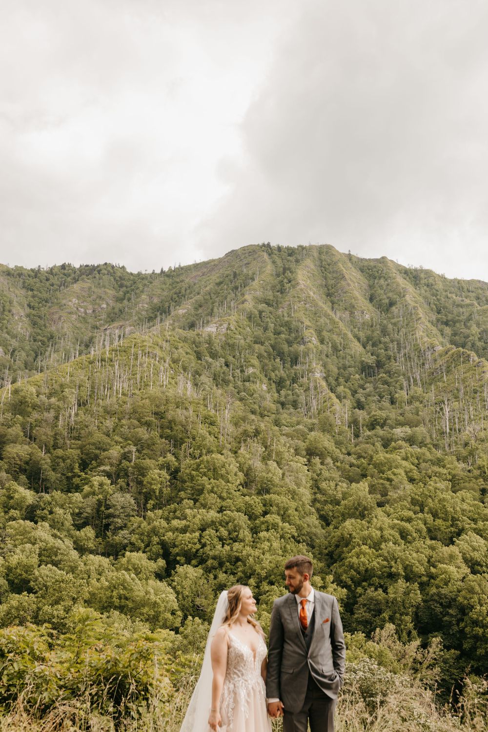 Bride and groom hold hands while standing before a dramatic green mountain landscape on their wedding day.