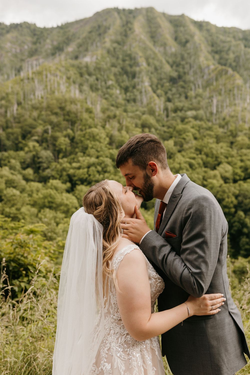 Groom in gray suit kisses bride in lace wedding gown on a scenic mountain overlook.