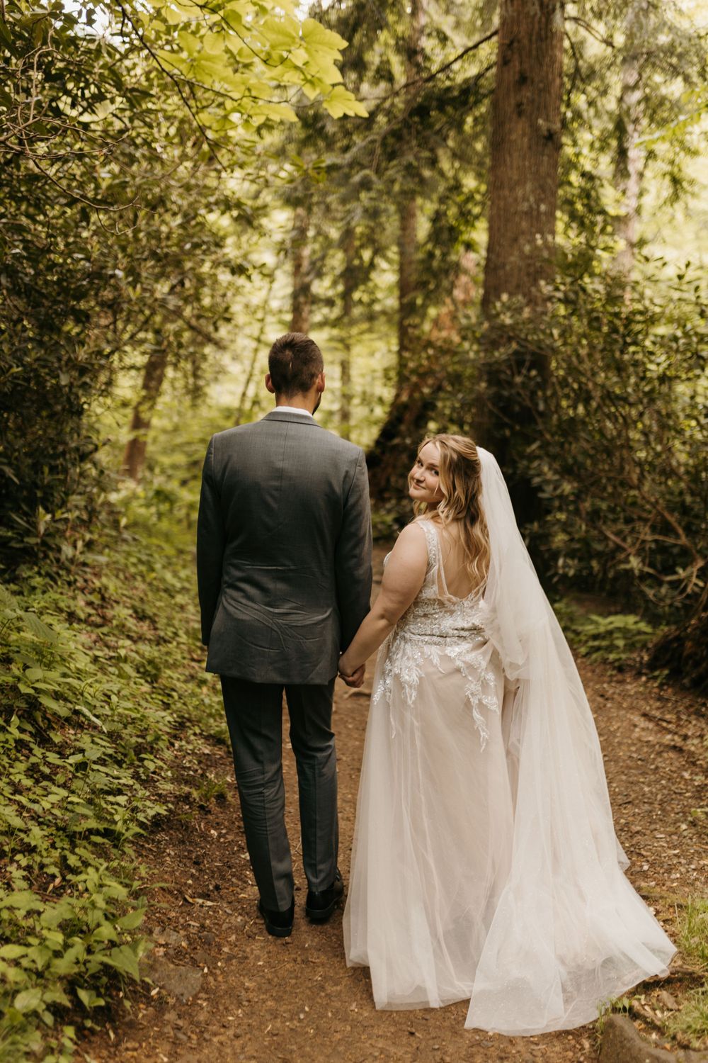 Bride glances back smiling while holding groom's hand walking along a lush green forest trail.