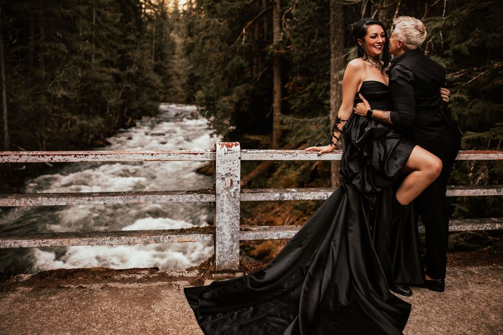 Dramatic photo of couple in black formal attire posing by a wooden railing with flowing dress in a forest setting.