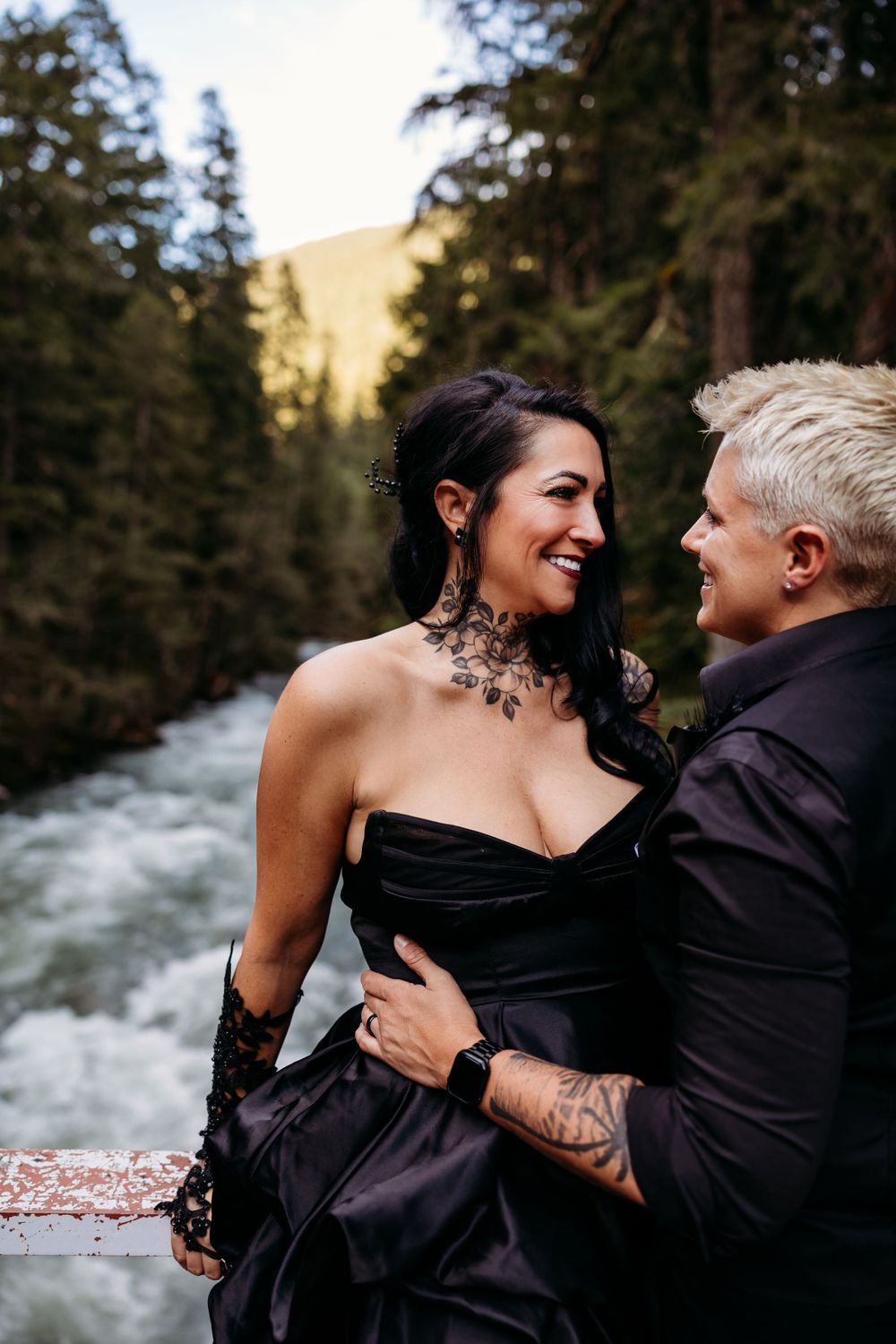 A romantic couple embraces by a rushing river in the forest wearing black formal attire.