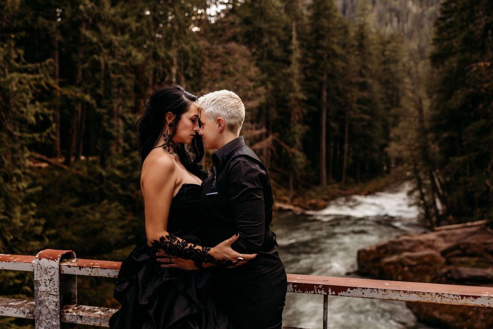A couple in black attire embraces on a bridge over a rushing river in autumn.