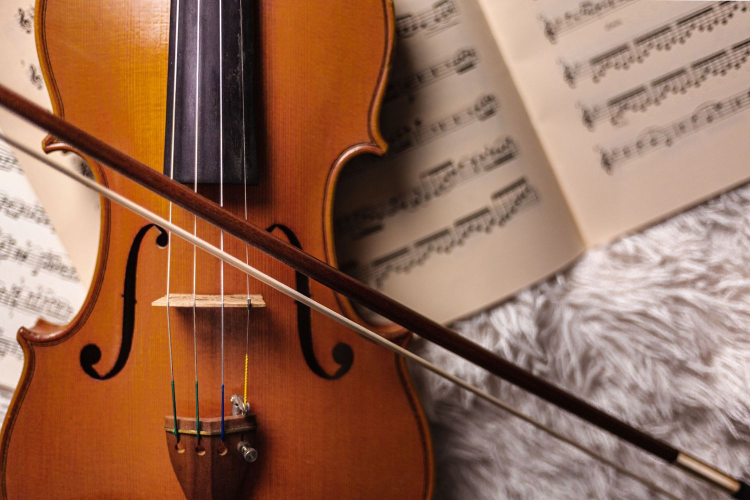 Close-up of a violin with a bow resting on its strings, placed on a fuzzy surface with an open sheet of music