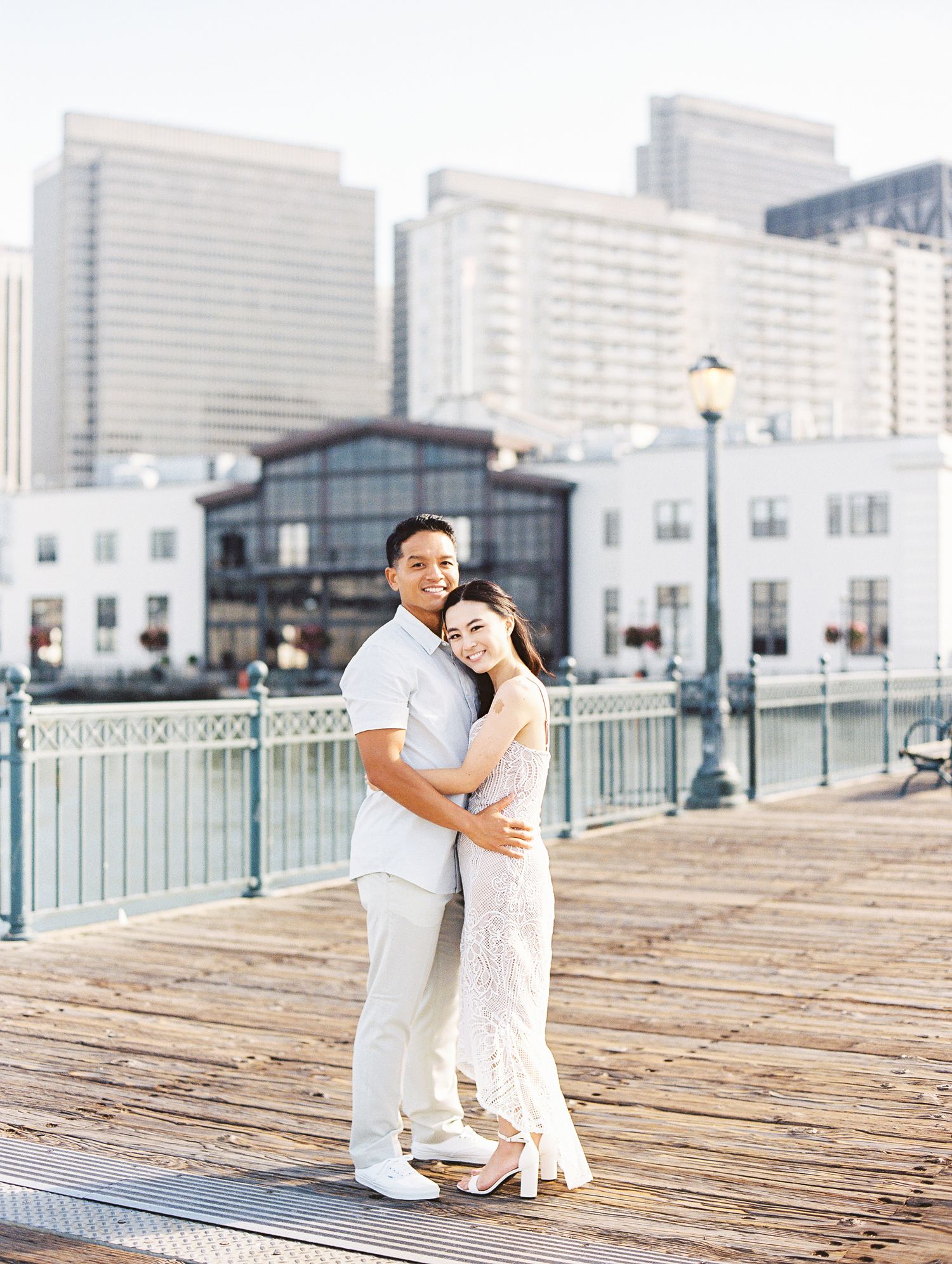 A couple in white outfits embracing on a waterfront boardwalk with modern buildings behind them.