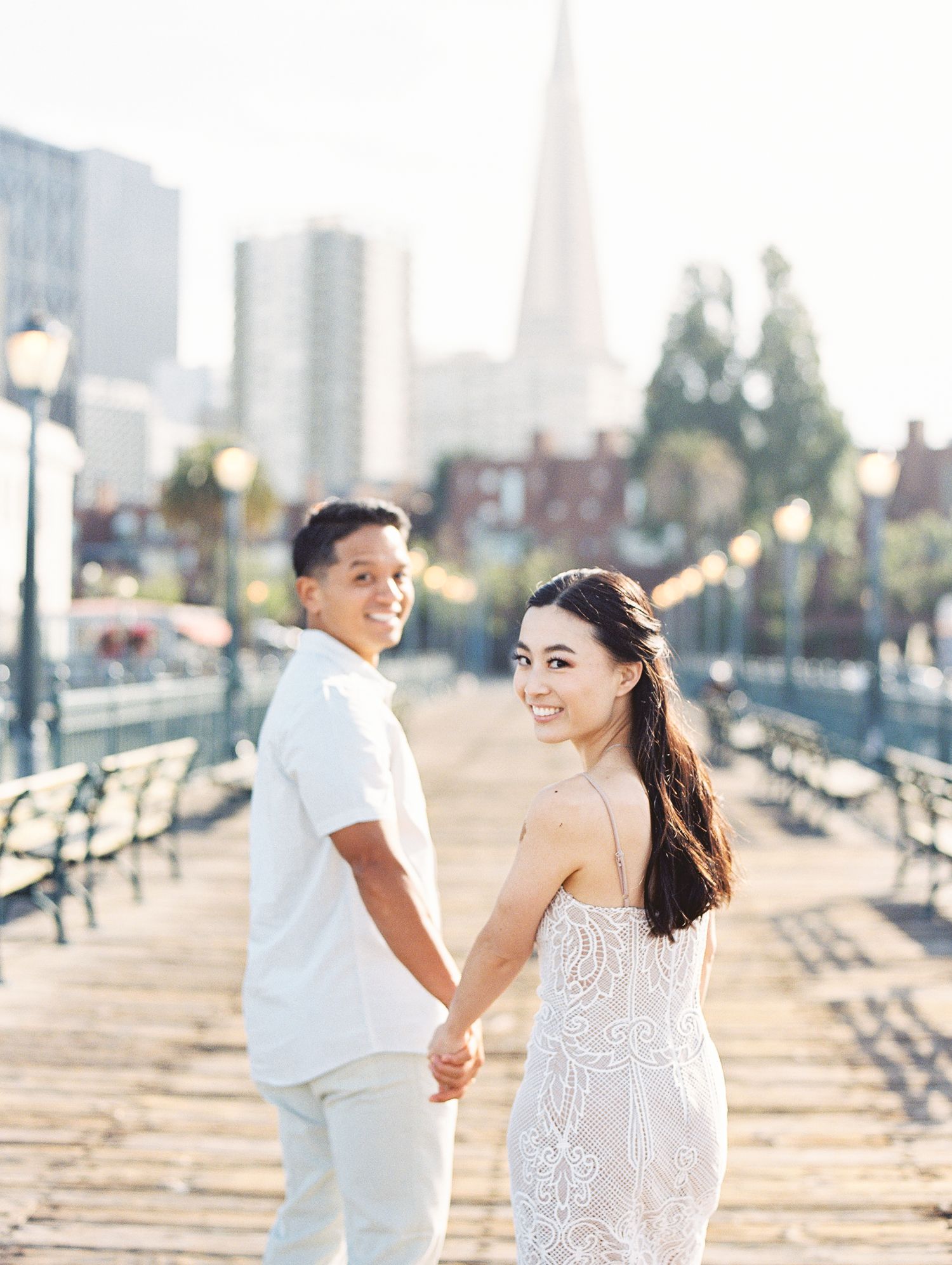 A couple in white outfits holding hands on a wooden pier with San Francisco's skyline behind them.