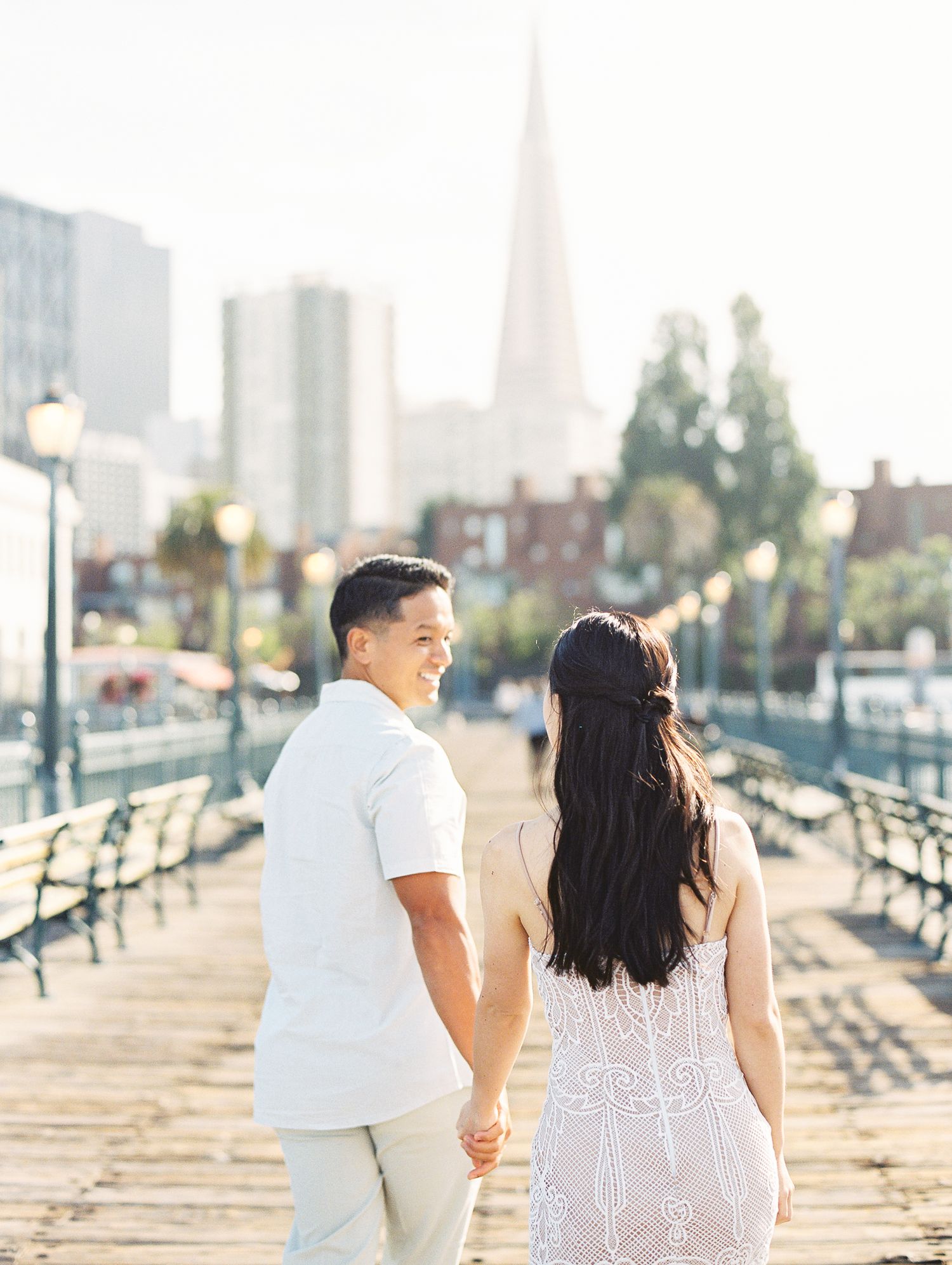 A couple walking hand in hand on a pier with the city skyline and palm trees in the background.