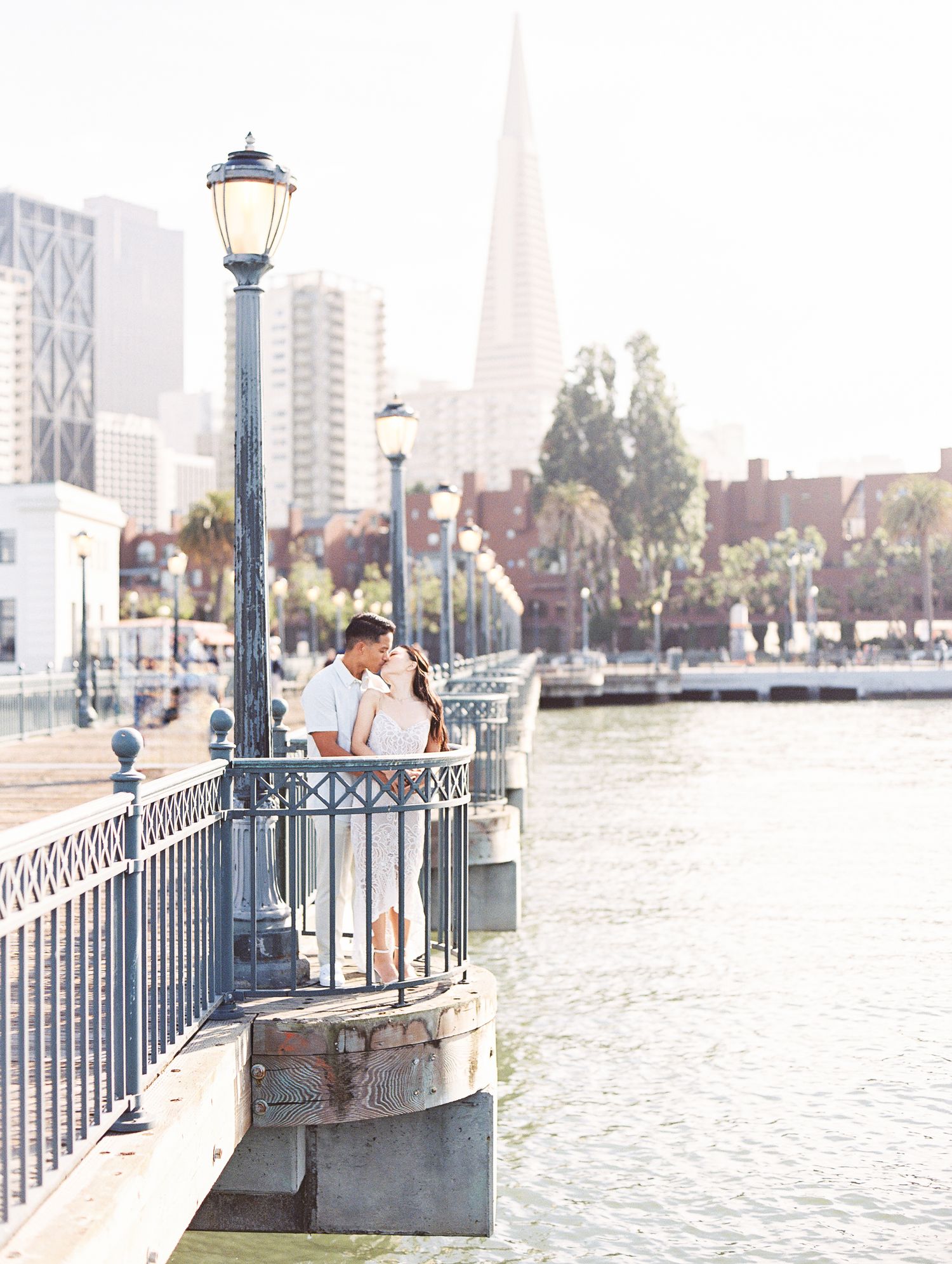 A couple standing by a vintage lamp post on a pier with the Transamerica Pyramid visible in the distance.