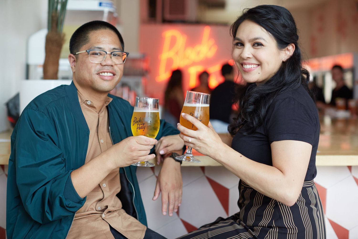 Couple toasting with glasses of beer at a restaurant with neon lights in background.