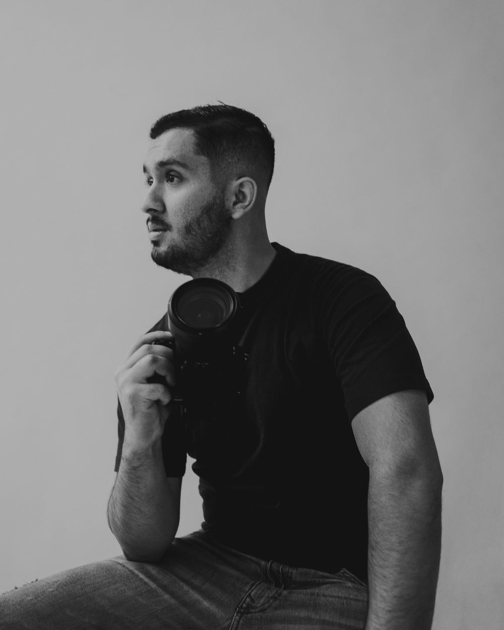 A man in a black t-shirt sits holding headphones against a neutral background in a black and white portrait.