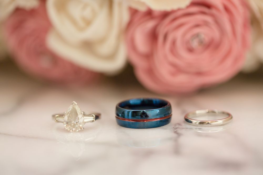 Three wedding rings arranged on white fabric including a blue band and two silver bands with roses in background.